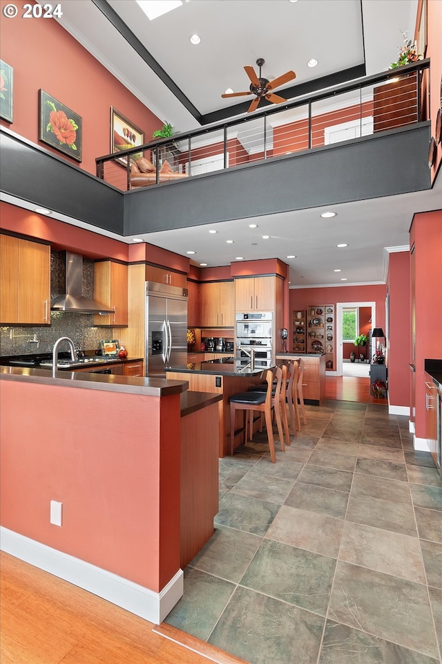 kitchen with wall chimney range hood, ceiling fan, backsplash, appliances with stainless steel finishes, and dark countertops