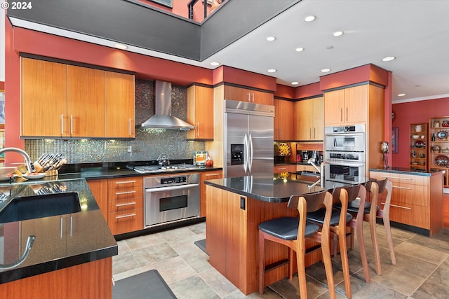kitchen with a sink, wall chimney exhaust hood, stainless steel appliances, a kitchen bar, and backsplash
