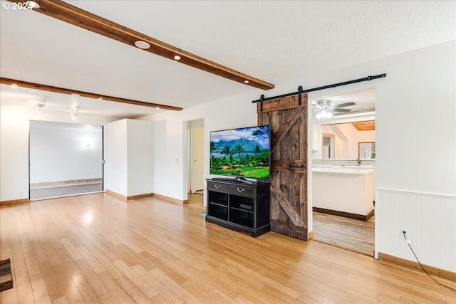 unfurnished living room featuring ceiling fan, light hardwood / wood-style floors, beamed ceiling, and a barn door