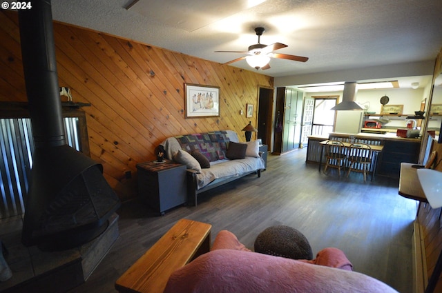 living room featuring a textured ceiling, ceiling fan, wooden walls, dark wood-type flooring, and a wood stove