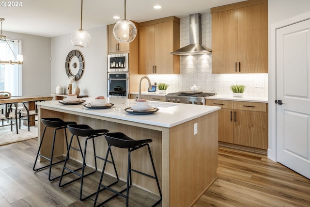 kitchen with a center island with sink, stainless steel appliances, light countertops, a sink, and wall chimney range hood