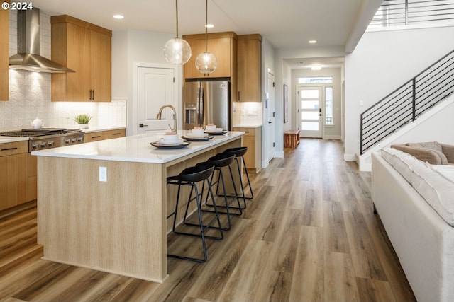 kitchen featuring an island with sink, wall chimney exhaust hood, a breakfast bar, stainless steel appliances, and light countertops