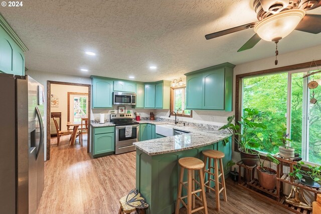 kitchen with kitchen peninsula, light stone counters, ceiling fan, light hardwood / wood-style floors, and appliances with stainless steel finishes