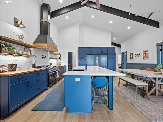 kitchen featuring wall chimney range hood, stainless steel appliances, decorative backsplash, a center island with sink, and blue cabinets