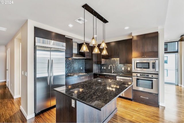 kitchen with built in appliances, a kitchen island, wall chimney range hood, tasteful backsplash, and hanging light fixtures