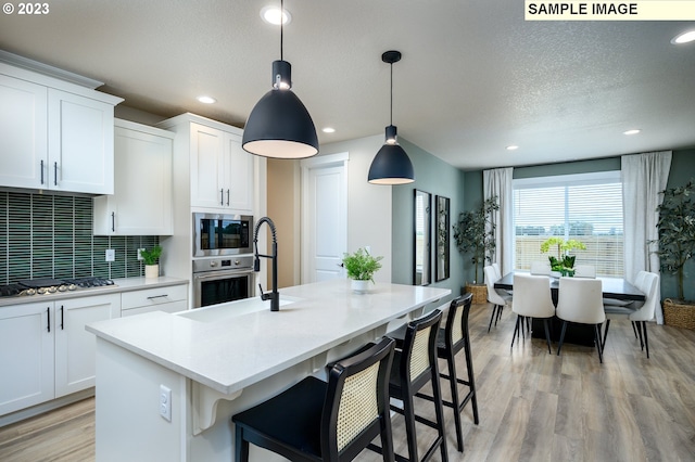 kitchen featuring a center island with sink, sink, appliances with stainless steel finishes, decorative light fixtures, and white cabinetry