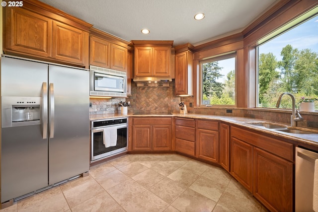 kitchen featuring decorative backsplash, under cabinet range hood, brown cabinets, a sink, and appliances with stainless steel finishes