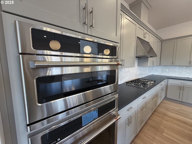 kitchen with stainless steel appliances, dark countertops, light wood-style flooring, backsplash, and under cabinet range hood