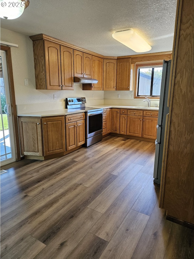 kitchen with under cabinet range hood, stainless steel electric range oven, freestanding refrigerator, brown cabinetry, and dark wood-style flooring