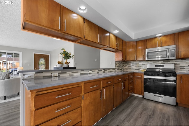 kitchen with brown cabinetry, dark wood finished floors, a peninsula, appliances with stainless steel finishes, and tasteful backsplash