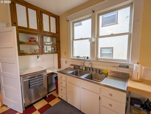 kitchen with stainless steel dishwasher, a sink, light floors, white cabinetry, and tile counters