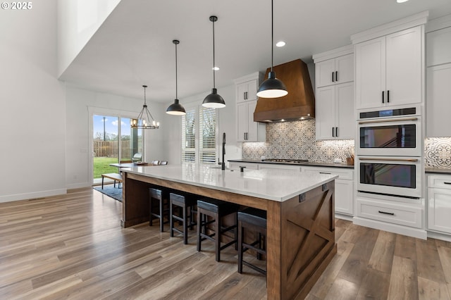 kitchen featuring premium range hood, white double oven, a center island with sink, and white cabinets