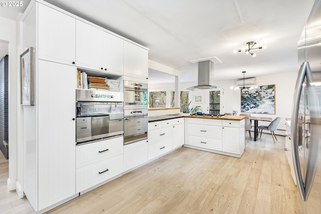 kitchen with island exhaust hood, a wall mounted AC, appliances with stainless steel finishes, light wood-type flooring, and a peninsula