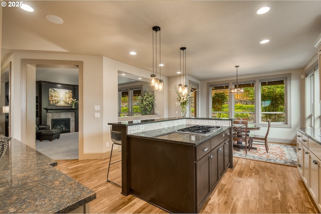 12483 SW Autumn View St, Tigard, OR 97224 - kitchen with stainless steel gas cooktop, light wood finished floors, a fireplace, dark brown cabinetry, and dark stone countertops