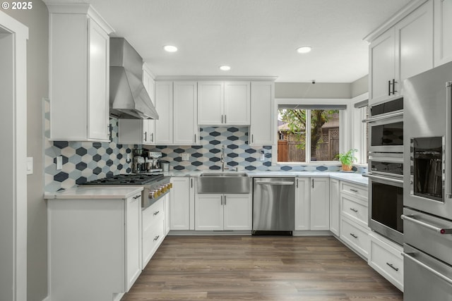 kitchen featuring a sink, stainless steel appliances, wall chimney range hood, light countertops, and white cabinetry