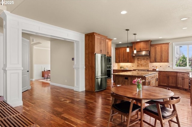 kitchen with under cabinet range hood, stainless steel appliances, backsplash, dark wood-type flooring, and decorative columns