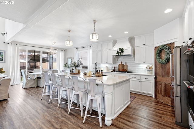 kitchen featuring sink, custom exhaust hood, a center island with sink, pendant lighting, and white cabinets