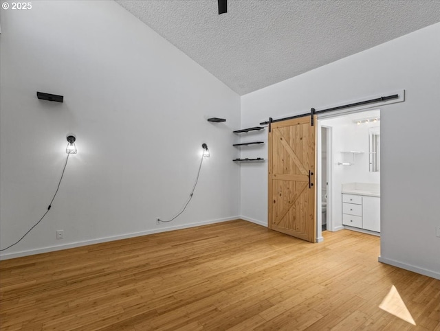 unfurnished bedroom featuring light wood-style flooring, a textured ceiling, a barn door, vaulted ceiling, and baseboards