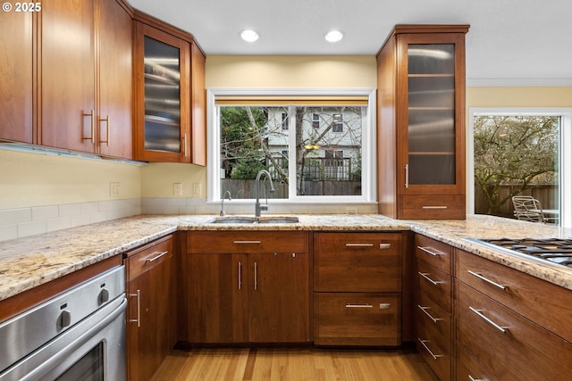 kitchen with light stone counters, light wood finished floors, stainless steel appliances, a sink, and glass insert cabinets