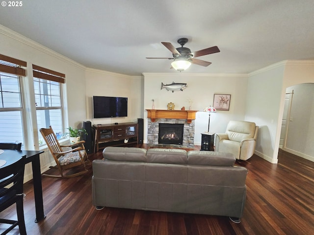 living room featuring dark wood-style flooring, crown molding, baseboards, a ceiling fan, and a stone fireplace