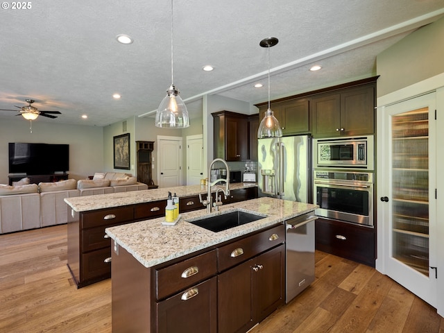 kitchen with dark brown cabinetry, an island with sink, a sink, light wood finished floors, and appliances with stainless steel finishes