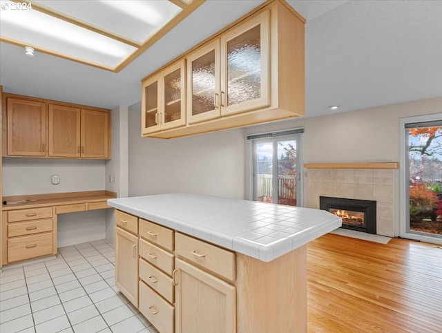 kitchen with light brown cabinets, a tile fireplace, built in desk, tile counters, and a kitchen island