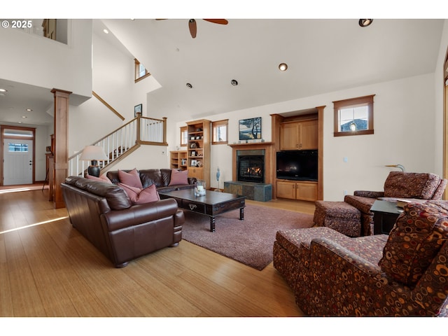 living area with a ceiling fan, a fireplace, light wood-style floors, a high ceiling, and stairs