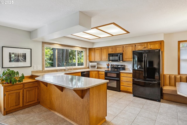 kitchen with brown cabinetry, a sink, light tile patterned floors, black appliances, and a peninsula