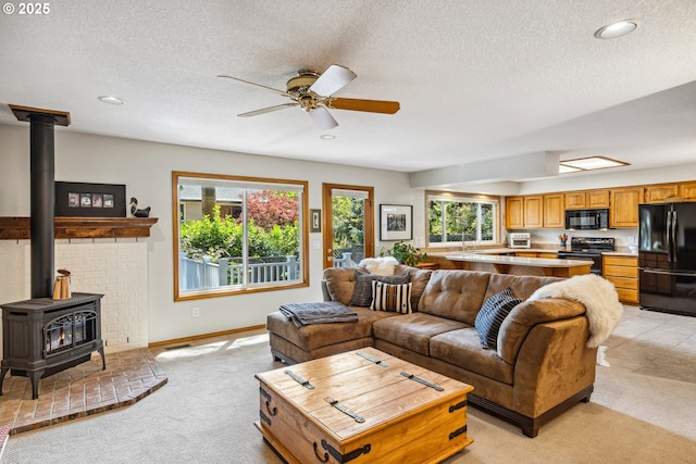 living room featuring a wood stove, baseboards, ceiling fan, light colored carpet, and a textured ceiling