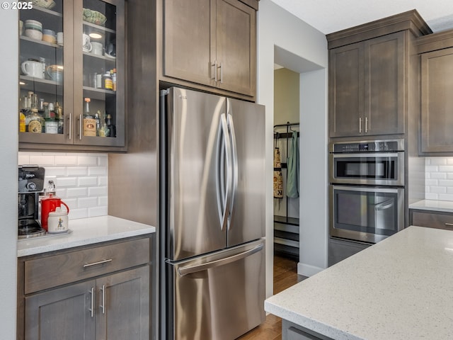 kitchen featuring backsplash, appliances with stainless steel finishes, glass insert cabinets, light stone countertops, and dark brown cabinets