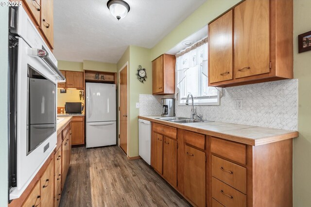 kitchen with brown cabinetry, decorative backsplash, dark wood-style floors, white appliances, and a sink