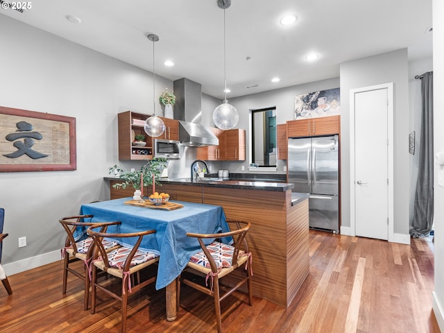 kitchen with kitchen peninsula, pendant lighting, appliances with stainless steel finishes, wall chimney exhaust hood, and hardwood / wood-style flooring