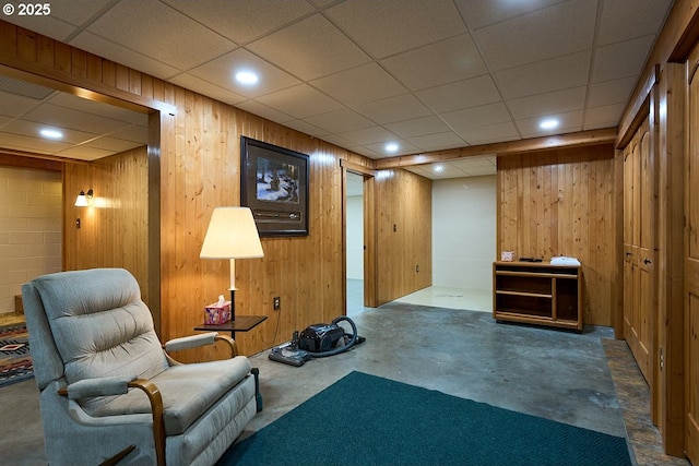 sitting room featuring wooden walls, concrete flooring, and a drop ceiling