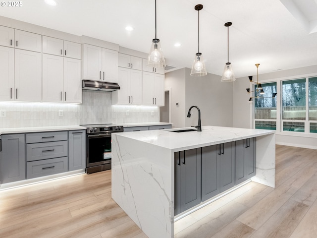 kitchen with under cabinet range hood, a sink, hanging light fixtures, stainless steel electric stove, and an island with sink