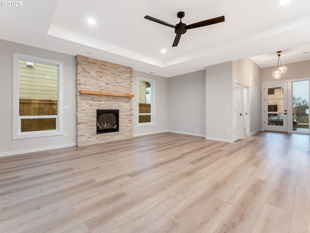 unfurnished living room with light wood-style flooring, a tray ceiling, baseboards, and a stone fireplace