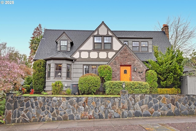 tudor-style house with stucco siding, stone siding, cooling unit, a shingled roof, and a chimney