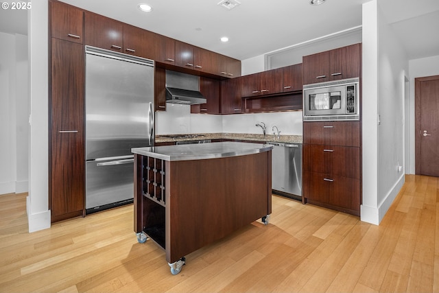 kitchen with ventilation hood, built in appliances, dark brown cabinets, a kitchen island, and light wood-type flooring