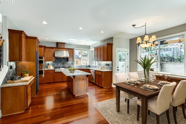14702 SE Badger Creek Rd, Happy Valley, OR 97086 - kitchen featuring wall chimney exhaust hood, stainless steel appliances, brown cabinetry, decorative backsplash, and dark wood-style floors