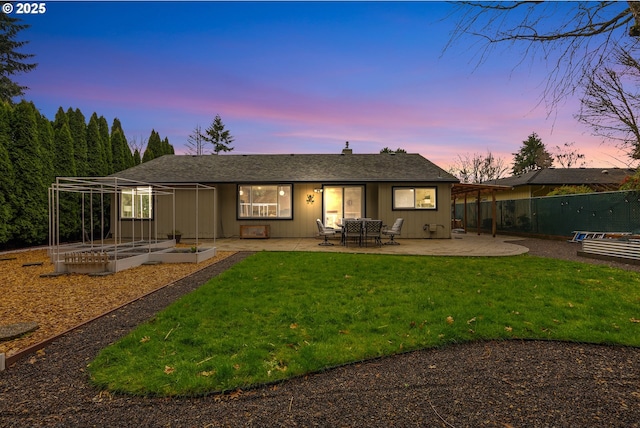 back of house at dusk with a garden, fence, a patio, roof with shingles, and a lawn