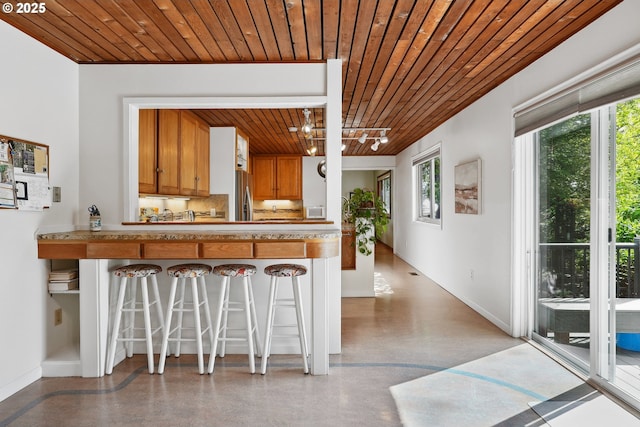kitchen featuring a breakfast bar area, brown cabinetry, stainless steel refrigerator with ice dispenser, wooden ceiling, and baseboards
