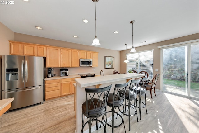 kitchen featuring light brown cabinetry, hanging light fixtures, appliances with stainless steel finishes, light wood-type flooring, and an island with sink