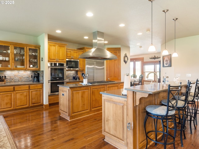 kitchen featuring an island with sink, stainless steel appliances, island range hood, dark wood-type flooring, and backsplash