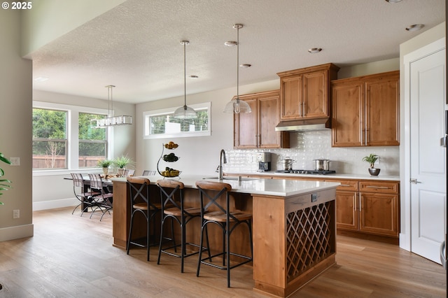 kitchen with a center island with sink, light countertops, brown cabinetry, a sink, and under cabinet range hood