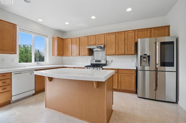 kitchen featuring brown cabinetry, a kitchen island, under cabinet range hood, tile counters, and stainless steel appliances