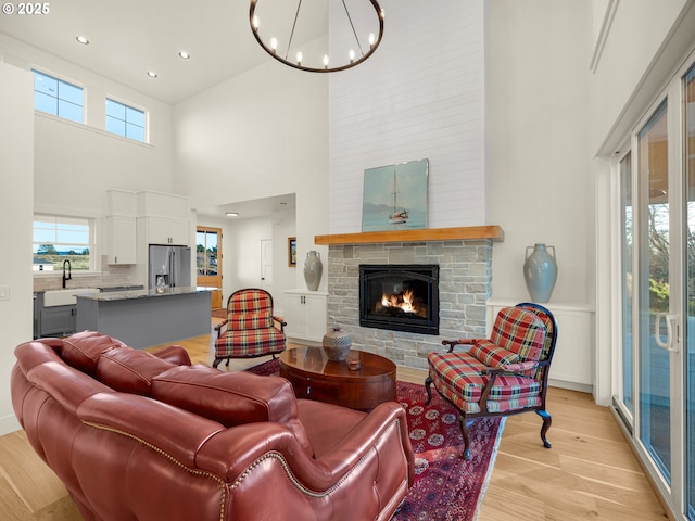 living room featuring a stone fireplace, sink, an inviting chandelier, light wood-type flooring, and a high ceiling