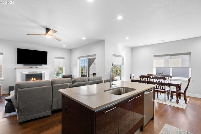kitchen with dark wood-style flooring, dark brown cabinets, a sink, a warm lit fireplace, and an island with sink