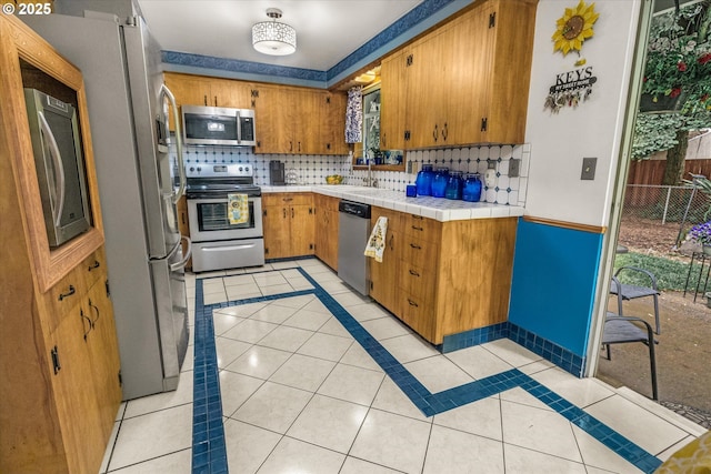 kitchen featuring stainless steel appliances, light tile patterned floors, backsplash, brown cabinetry, and a sink