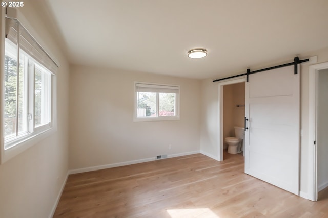 unfurnished bedroom featuring ensuite bath, a barn door, light wood finished floors, baseboards, and visible vents