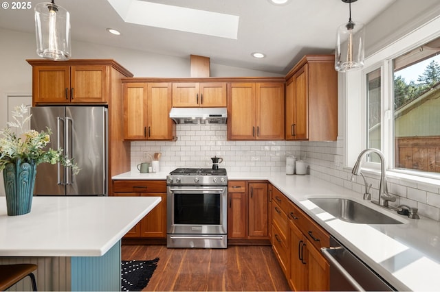 kitchen with under cabinet range hood, lofted ceiling with skylight, appliances with stainless steel finishes, brown cabinetry, and a sink