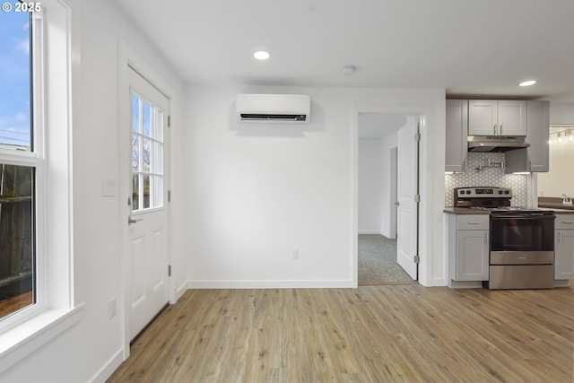 kitchen featuring a wall unit AC, tasteful backsplash, light wood-style flooring, stainless steel range with electric stovetop, and under cabinet range hood